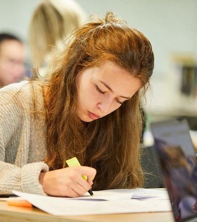 A student is focused on writing while studying in a busy environment.