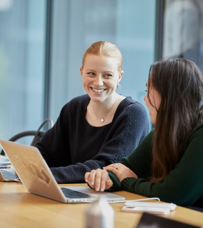 Two women are engaging in a friendly discussion while sitting at a table with laptops in a modern office environment.