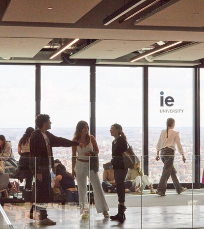 Students and staff engaging in casual conversation inside a modern university building with large windows overlooking a city.