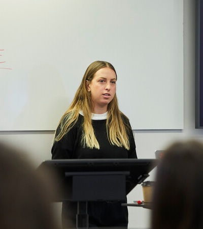 A woman is giving a presentation in a classroom with a Q&A slide displayed behind her.