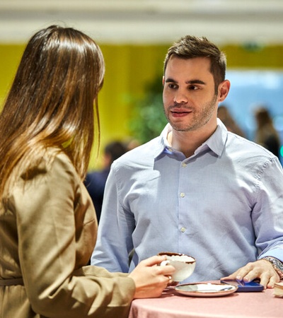 A man and a woman are engaged in a conversation over coffee in a busy cafe setting.