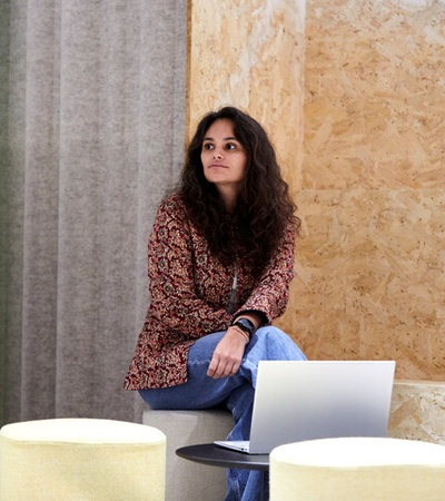 A woman sits on a stool in a modern decorated room with a laptop on a small round table, looking contemplative.