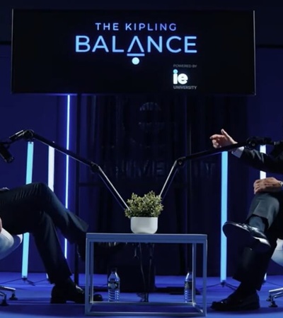 Two men in business attire are sitting across from each other in a modern interview setup with a blue aesthetic, featuring a sign that reads 'The Kipling Balance'.