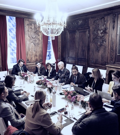 A formal meeting in an elegant room with a long table and attendees engaged in discussion.