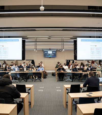 A group of people seated in a modern conference room with presentations displayed on screens.