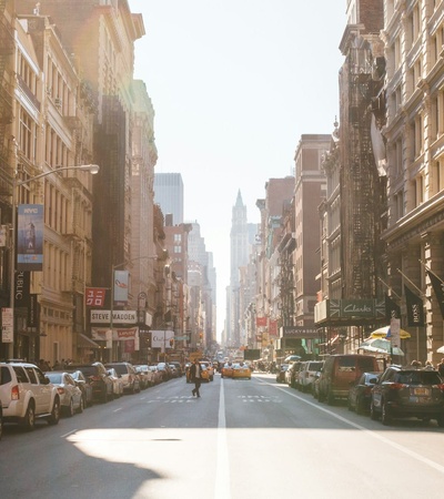 A sunlit urban street scene with cars parked and people walking in a busy downtown area.