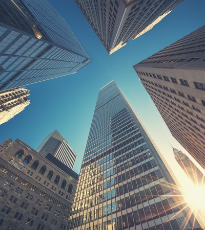 Looking up at towering skyscrapers against a clear blue sky with sunlight shining through.