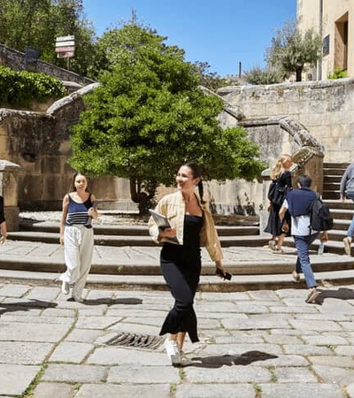 People walking through a historic stone staircase surrounded by aged buildings and trees under a clear sky.