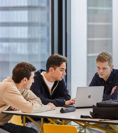 Three young men are collaborating on a laptop at a modern workspace with a city view.