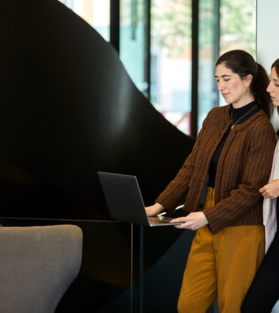 Two women standing and using a laptop together in a modern office setting.