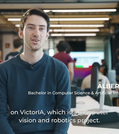 A young man stands in an office environment, speaking about a project named VictorIA, related to computer vision and robotics.