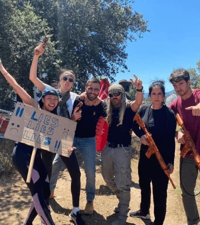 A group of seven people posing outdoors, some holding props like a sign 'II HS CLUB 25' and musical instruments, expressing joy and enthusiasm.