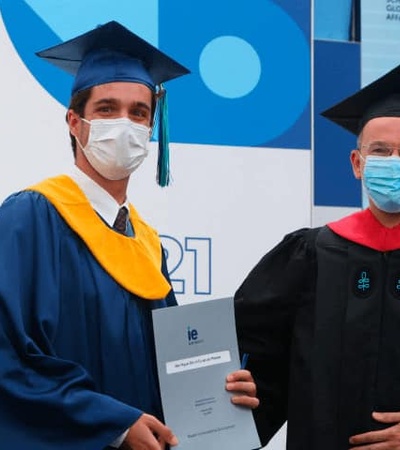 A young graduate in a blue and gold cap and gown receives a diploma from a professor wearing a red and black academic robe, both wearing protective face masks.