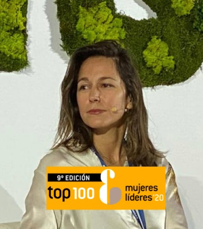 A woman sits on stage at an event for 'Top 100 Women Leaders 2020' with a green plant background.