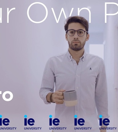 A man named Alvaro standing in an office setting holding a coffee mug, with text overlay 'Your Own Path' and multiple IE University logos.