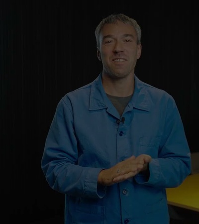 A man in a blue shirt standing in a modern office environment, smiling at the camera.