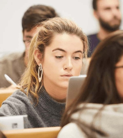 A focused young woman using a laptop in a classroom full of students.
