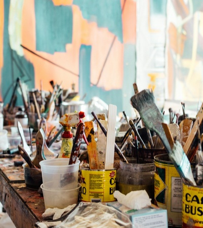A cluttered artist's table filled with paint brushes, open paint cans, and various painting supplies in front of a colorful abstract painting.