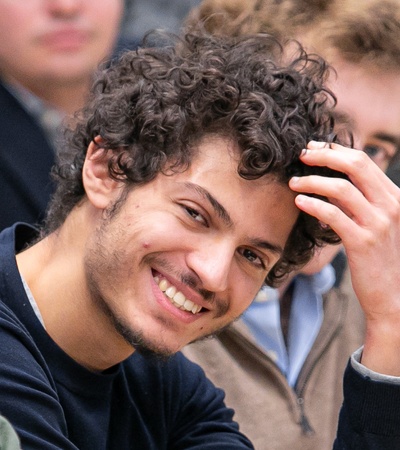 A young man with curly hair smiling and adjusting his hair in a public setting