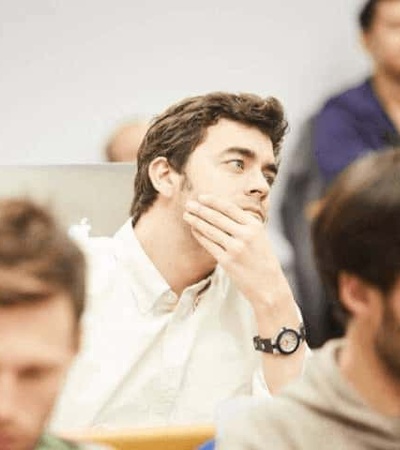 A young man in a lecture hall looks thoughtful while resting his chin on his hand.