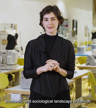 A person presenting in a fashion design studio with sewing machines in the background.