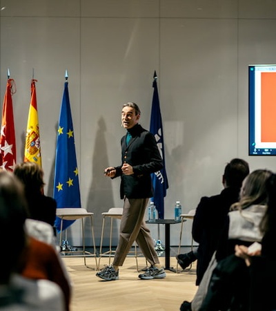 A person presenting at a conference with flags of Spain, the European Union, and another in the background.