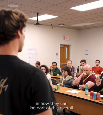 A young man is speaking to a diverse group of students in a classroom setting, while the group appears focused and engaged.