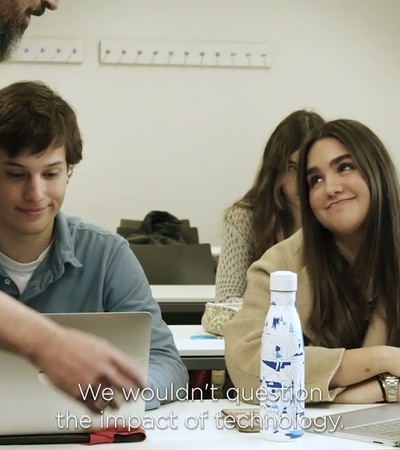 A classroom scene with students engaged in a discussion while using laptops.