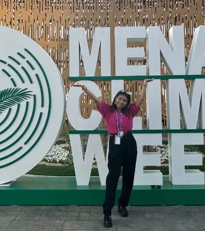 A person posing in front of a large sign that reads 'MENA Climate Week' with a circular logo featuring a palm design.