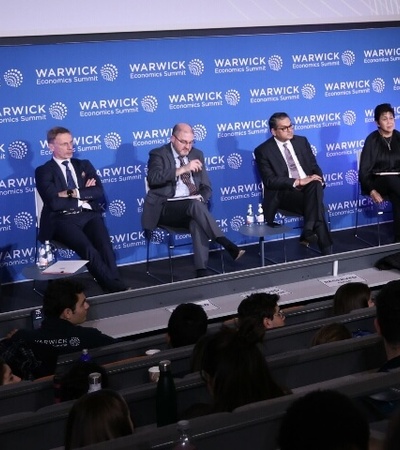 A panel of six people, consisting of five men and one woman, seated on stage in an auditorium with a 'Warwick' banner in the background.