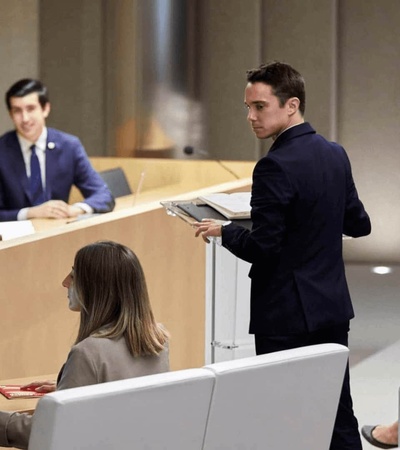 A man is speaking at a podium in a modern conference room with several attendees and a panel of two men and a woman.