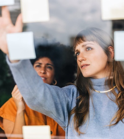 Two women collaborating on ideas using sticky notes on a glass window in an office setting.