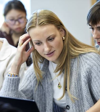 Two women and a man are focused on work at a cluttered office desk with one woman speaking on the phone.