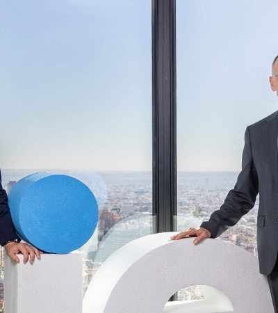 Two businessmen in suits standing next to a large blue and white circular logo inside an office with a cityscape visible through the window.