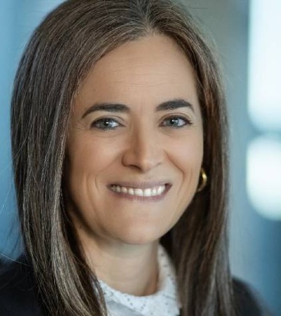 Portrait of a smiling middle-aged woman with medium-length brown hair, wearing a black jacket and a white blouse in an office setting.