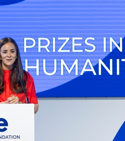 A woman in a red top stands at a podium, presenting at an event titled 'PRIZES IN THE HUMANITIES' organized by the IE Foundation.