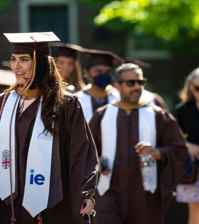 A group of graduates in brown and white robes walking during a sunny graduation ceremony