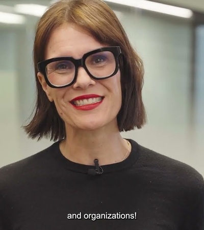 A woman with glasses smiling in an office environment with subtitles displayed.