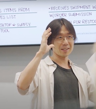 A man with glasses gestures while speaking in front of a whiteboard during a meeting.