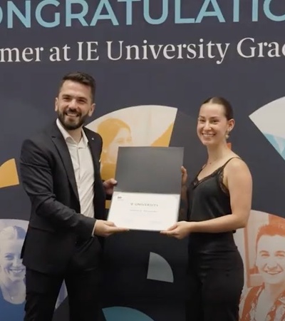 A man and a woman smiling while holding a diploma in front of a congratulatory banner at IE University.