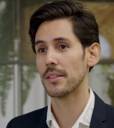 A man with a light beard and mustache, wearing a dark blazer over a white shirt, stands in an indoor setting with blurred green plants in the background.