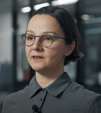 A woman in a gray shirt is speaking, wearing a lavalier microphone, in an office setting with blurred background.