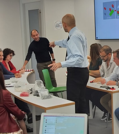 A group of adults engaged in a discussion at a workshop with one man standing and pointing at a screen displaying graphs.