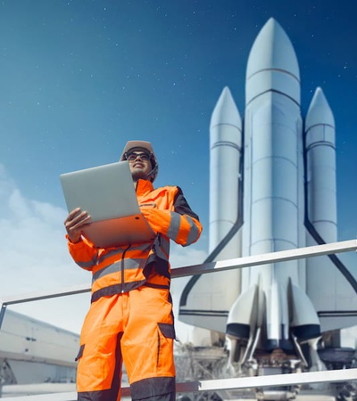 A technician in an orange uniform is using a laptop while standing near a large rocket.