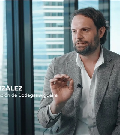 A man in a gray blazer gestures while speaking in an office environment with glass walls and city buildings visible in the background.