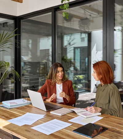 Two women are having a discussion at a wooden table in a modern office space with plants and glass windows.