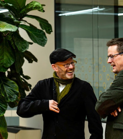 Two men in a casual business setting, engaging in a cheerful conversation near a large potted plant.