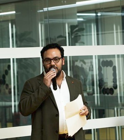 A man with glasses and a beard is speaking into a microphone in an office setting with other seated people listening.