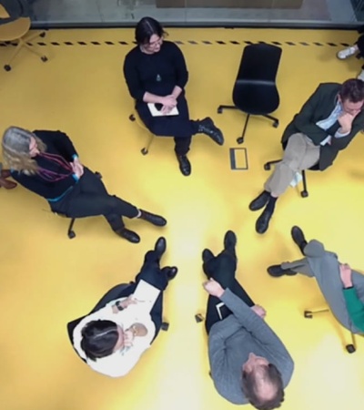 An overhead view of a group of people sitting and standing on a yellow floor, some conversing and others using smartphones.