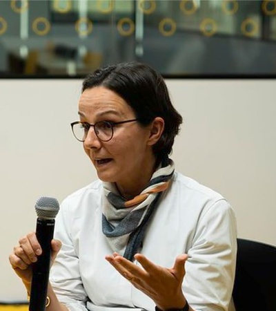 A woman is speaking into a microphone while gesturing with her hands during an indoor event.